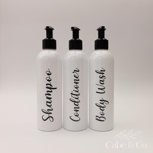 May include: Three white cylindrical plastic bottles with black pump dispensers. Each bottle features black cursive text printed vertically on the front, reading Shampoo, Conditioner, and Body Wash. The bottles are arranged in a row.