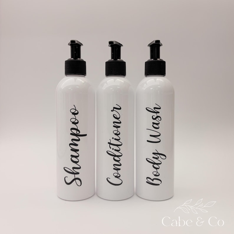 May include: Three white cylindrical plastic bottles with black pump dispensers. Each bottle features black cursive text printed vertically on the front, reading Shampoo, Conditioner, and Body Wash. The bottles are arranged in a row.