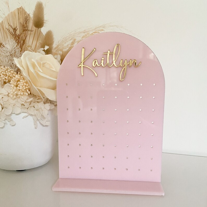 May include: A pink acrylic earring display stand with an arched top and a rectangular base. The stand has multiple holes for earrings and features the name "Kaitlyn" in gold script. A floral arrangement is in the background.