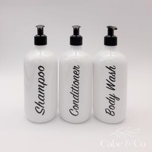 May include: Three white cylindrical plastic bottles with black pump dispensers. Each bottle features black cursive text printed vertically on the front, reading Shampoo, Conditioner, and Body Wash respectively. The bottles have a smooth finish.