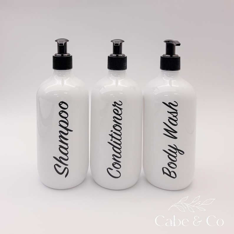May include: Three white cylindrical plastic bottles with black pump dispensers. Each bottle features black cursive text printed vertically on the front, reading Shampoo, Conditioner, and Body Wash respectively. The bottles have a smooth finish.