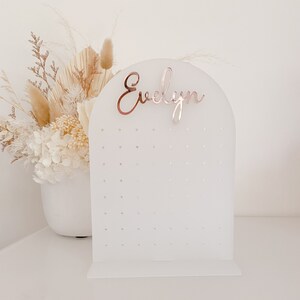 May include: A white acrylic earring display stand with an arched top and a base. The stand has multiple small holes for earrings and a rose gold name, "Evelyn", at the top. A bouquet of dried flowers is in the background.