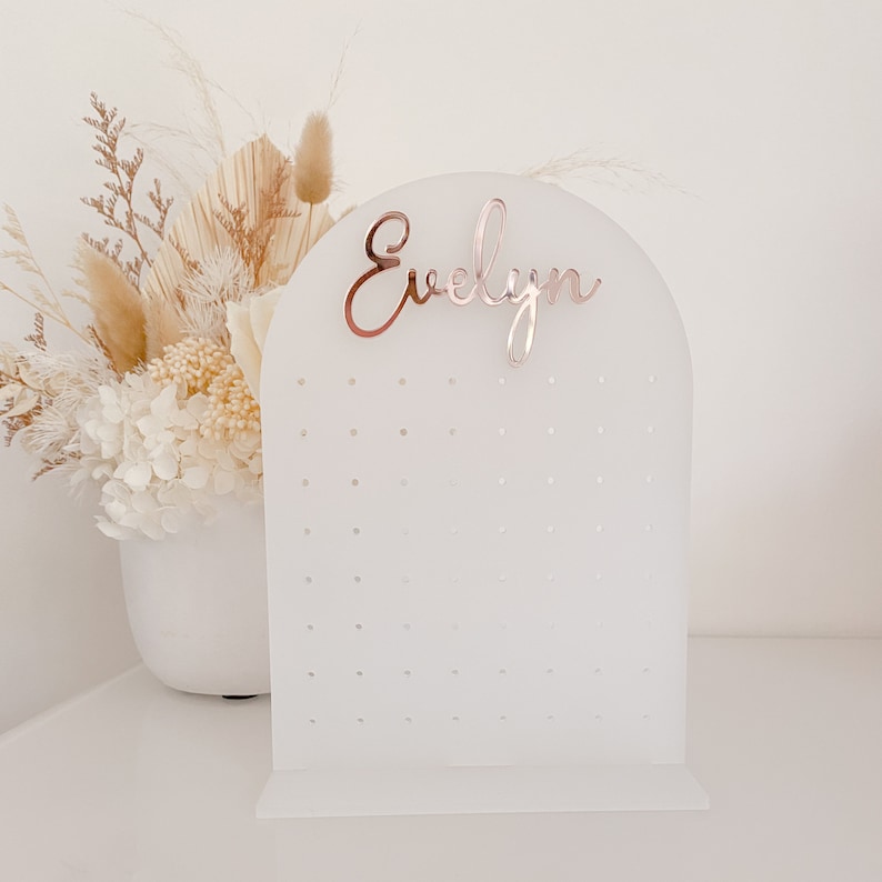 May include: A white acrylic earring display stand with an arched top and a base. The stand has multiple small holes for earrings and a rose gold name, "Evelyn", at the top. A bouquet of dried flowers is in the background.