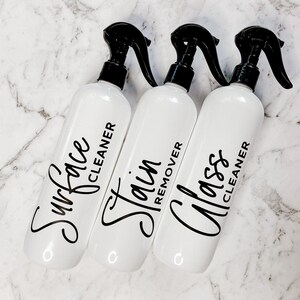 May include: Three white spray bottles with black trigger tops and labels. The labels read "Surface Cleaner", "Stain Remover", and "Glass Cleaner".