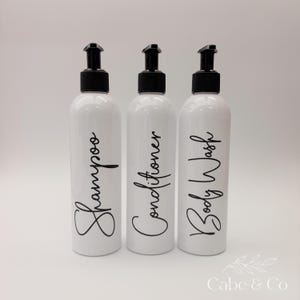 May include: Three white cylindrical plastic bottles with black pump dispensers. Each bottle features black cursive text printed on the front, reading Shampoo, Conditioner, and Body Wash. The bottles are arranged in a row against a plain background.