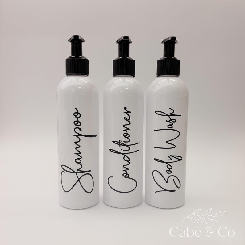 May include: Three white cylindrical plastic bottles with black pump dispensers. Each bottle features black cursive text printed on the front, reading Shampoo, Conditioner, and Body Wash. The bottles are arranged in a row against a plain background.