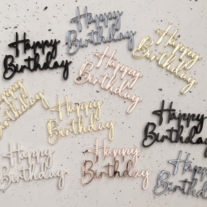 May include: Assortment of acrylic cake toppers in various colours, including gold, silver, black, and white. Each topper features the phrase "Happy Birthday" in a cursive script.