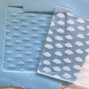 May include: A clear acrylic stamp with a repeating pattern of white clouds. The stamp is placed on top of a light blue silicone sheet with the same cloud pattern embossed into the surface.