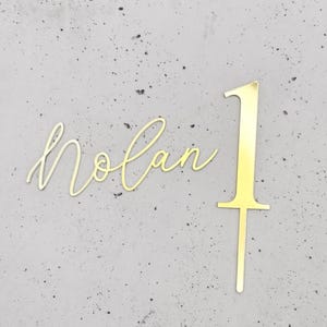 May include: A gold cake topper featuring the number "1" and the name "Nolan" in a cursive script. The number has a stake for insertion into a cake. The name is also in a cursive font, suitable for a birthday cake.