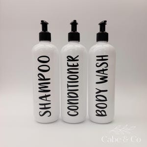 May include: Three white cylindrical plastic bottles with black pump dispensers. Each bottle features bold black lettering vertically printed on the front, labeled Shampoo, Conditioner, and Body Wash, providing a clear and organized storage solution.