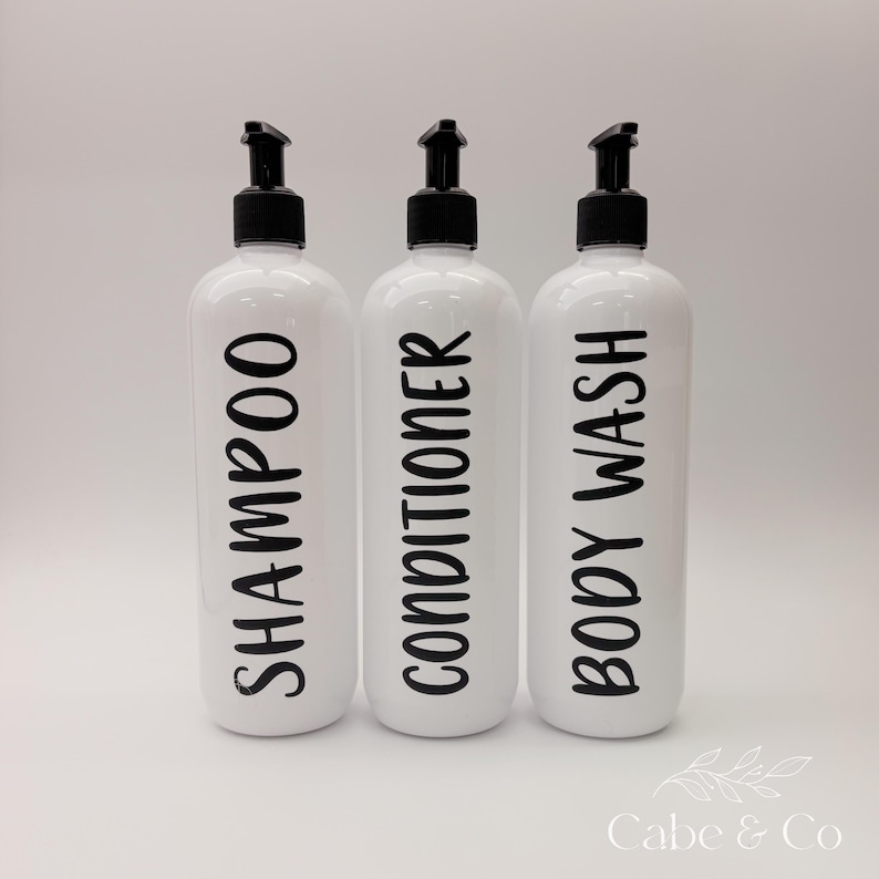 May include: Three white cylindrical plastic bottles with black pump dispensers. Each bottle features bold black lettering vertically printed on the front, labeled Shampoo, Conditioner, and Body Wash, providing a clear and organized storage solution.