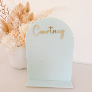 May include: A light blue earring display stand with the name "Courtney" in gold script. The stand has multiple small holes for holding earrings and a curved top. A white vase with dried flowers is in the background.