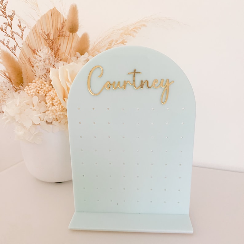 May include: A light blue earring display stand with the name "Courtney" in gold script. The stand has multiple small holes for holding earrings and a curved top. A white vase with dried flowers is in the background.