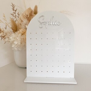 May include: A white acrylic earring display stand with an arched top and a base. The word "Sophie" is in silver script on the top. The display has multiple holes for earrings. A vase of dried flowers is in the background.