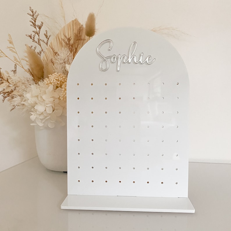 May include: A white acrylic earring display stand with an arched top and a base. The word "Sophie" is in silver script on the top. The display has multiple holes for earrings. A vase of dried flowers is in the background.