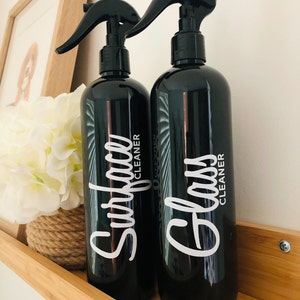 May include: Two black spray bottles with white labels. The bottle on the left says "Surface Cleaner" and the bottle on the right says "Glass Cleaner".