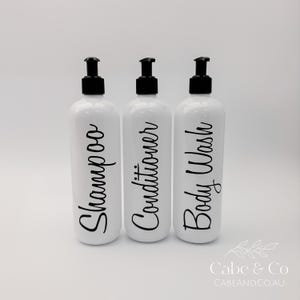 May include: Three white cylindrical plastic bottles with black pump dispensers. Each bottle features black cursive text printed vertically on the front, reading Shampoo, Conditioner, and Body Wash. The bottles are arranged in a row.