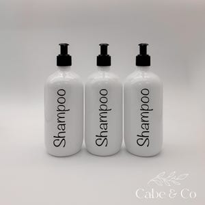 May include: Three white cylindrical bottles with black pump dispensers. Each bottle features the word Shampoo printed in a black, cursive-style font on the front. The bottles are arranged in a row against a plain, light-colored background.