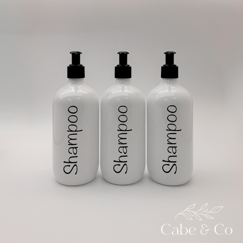 May include: Three white cylindrical bottles with black pump dispensers. Each bottle features the word Shampoo printed in a black, cursive-style font on the front. The bottles are arranged in a row against a plain, light-colored background.
