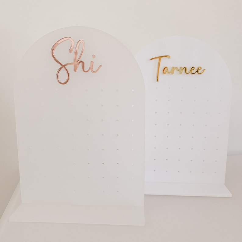 May include: Two white acrylic earring display stands. One stand has the name "Shi" in rose gold script, and the other has "Tarnce" in gold script. Each stand has a series of small holes for displaying earrings.
