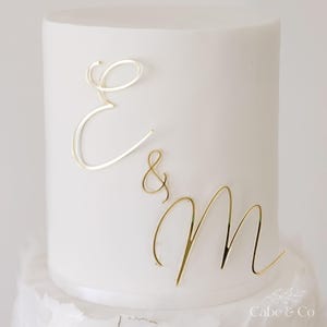 May include: A white cake decorated with gold cursive letters spelling "E & M". The cake has a smooth top and a textured base. The gold letters provide an elegant, decorative element to the cake's design.