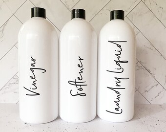 May include: Three white cylindrical plastic bottles with black screw-top caps. Each bottle features black cursive lettering on the front. The labels read Vinegar, Softener, and Laundry Liquid respectively, arranged in a row on a flat surface.