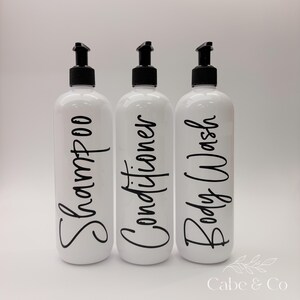 May include: Three white cylindrical plastic bottles with black pump dispensers. Each bottle features black cursive text printed vertically on the front, reading Shampoo, Conditioner, and Body Wash. The bottles are arranged in a single row.