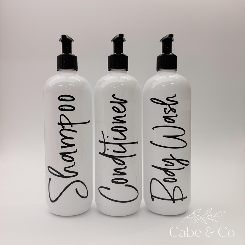 May include: Three white cylindrical plastic bottles with black pump dispensers. Each bottle features black cursive text printed vertically on the front, reading Shampoo, Conditioner, and Body Wash. The bottles are arranged in a single row.