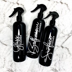 May include: Three black spray bottles with white labels and handwritten text. The labels read "Glass Cleaner", "Bathroom Cleaner", and "Surface Cleaner".