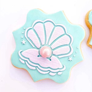 May include: A light blue sugar cookie decorated with a white seashell and a pink pearl. The cookie is shaped like a star.