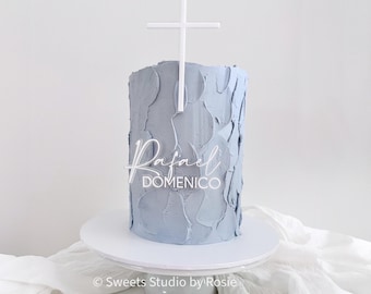 May include: A light blue cake with textured frosting, topped with a white cross and a clear acrylic nameplate that reads "Rafael Domenico". The cake sits on a white cake stand.