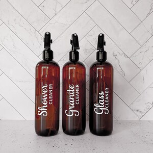 May include: Three amber glass spray bottles with black spray tops and white labels. The labels read "Shower Cleaner", "Granite Cleaner", and "Glass Cleaner".