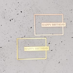 May include: Two metallic rectangular frames with the words "HAPPY BIRTHDAY" etched on a horizontal bar. One frame is gold, the other is rose gold. The frames are set against a speckled gray background.