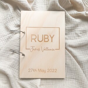 May include: A light wood baby memory book with a silver ring binder. The cover is engraved with the name "RUBY" and the text "James Vallance" and the date "27th May 2022".