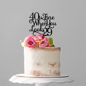 May include: A frosted cake with a black cake topper that reads "40 is fine when you look 29." The cake is decorated with pink flowers and macarons. The cake sits on a white cake stand. The cake is approximately 15 cm in height.