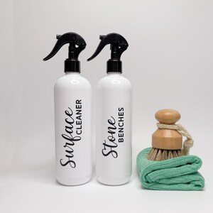 May include: Two white spray bottles with black trigger sprayers and labels. The bottle on the left says "Surface Cleaner" and the bottle on the right says "Stone Benches". A green cleaning cloth and a wooden brush are in the foreground.