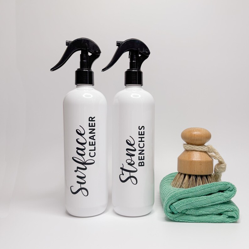 May include: Two white spray bottles with black trigger sprayers and labels. The bottle on the left says "Surface Cleaner" and the bottle on the right says "Stone Benches". A green cleaning cloth and a wooden brush are in the foreground.