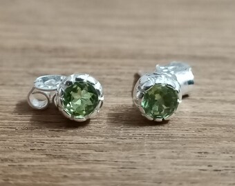 Könnte beinhalten: Ein Paar Ohrstecker mit runden, facettierten, hellgrünen Peridot-Edelsteinen in gewellten Silberfassungen. Jeder Ohrring verfügt über einen passenden Schmetterlingsverschluss aus Silber und besticht durch ein klassisches Design mit polierter Oberfläche.