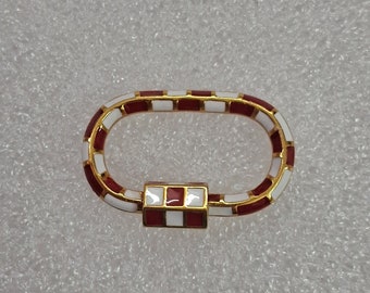 May include: A gold-toned metal clasp with a red and white enamel design. The clasp is oval-shaped and has a square-shaped piece in the center.