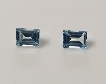 Op de afbeelding: Twee rechthoekige edelstenen in emerald-slijpvorm met een lichtblauwe kleur en hoge helderheid. Elke steen heeft een gefacetteerd oppervlak dat licht reflecteert en een transparante, ijsblauwe tint laat zien.