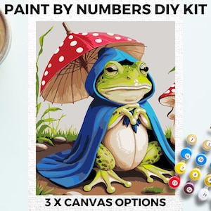 May include: Paint by numbers DIY kit featuring a green frog wearing a blue hooded cloak and holding a red and white mushroom umbrella. The kit includes numbered paints and a canvas.