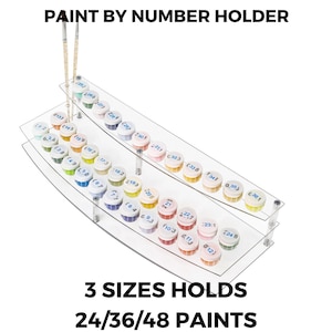 Puede incluir: Soporte transparente de acrílico para pintar por números con tres niveles, con capacidad para 24, 36 o 48 botes de pintura. El soporte tiene el texto "PAINT BY NUMBER HOLDER" y "3 SIZES HOLDS 24/36/48 PAINTS". Pinceles colocados encima.