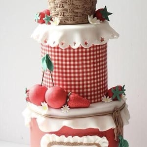 Può includere: Una torta a tre piani decorata con ciliegie rosse, fragole e un cestino intrecciato. Il piano centrale ha un motivo a scacchi rossi e bianchi. Il piano inferiore presenta il nome "Ginevra" in corsivo. La torta è adornata con fiori bianchi.