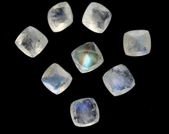 May include: Eight square-cut moonstone gemstones are arranged against a black background. The stones have a translucent, milky appearance with flashes of blue and iridescent color. The gemstones are likely for jewelry making.