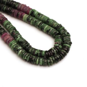 May include: Close-up of a necklace made of small, flat, round beads. The beads are a mix of green, black, and maroon colors. The necklace is on a white background. The beads appear to be made of stone or a similar material.