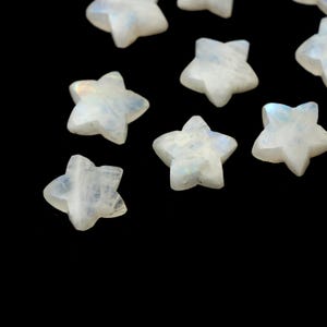 May include: A collection of small, star-shaped gemstones. The stones are a milky white color with iridescent flashes of blue and purple. The stars are scattered against a black background, creating a celestial effect. These gemstones are ideal for jewelry making or craft projects.