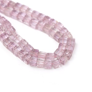 Puede incluir: Un collar de cuentas cúbicas de color púrpura claro. Las cuentas están ensartadas, creando una línea curva sobre un fondo blanco. Las cuentas tienen una cualidad translúcida, con sutiles variaciones de color.