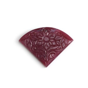 Könnte beinhalten: Ein fächerförmiger, tiefroter Edelstein mit aufwendigen Blumenschnitzereien. Das geschnitzte Design zeigt eine zentrale Blume, umgeben von detaillierten Blättern und wirbelnden Mustern. Der Edelstein ist vor weißem Hintergrund.