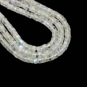 May include: Strands of translucent, square-cut moonstone beads. The beads have a subtle, iridescent sheen with flashes of blue and white. The beads are strung together, creating a flowing, organic shape against a black background. Ideal for jewelry making.