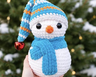May include: A crocheted snowman plush toy with a white body, a blue scarf, and a floppy blue, white, and orange striped hat. The snowman features black plastic eyes, an orange nose, and a small red bell attached to the tip of the hat.